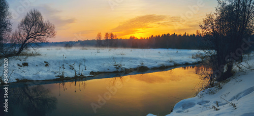 Frosty foggy winter panoramic landscape with small forest river and rising sun.