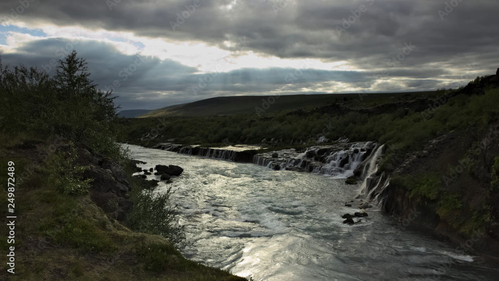 Longer exposure of Hraunfossar waterfall on Hvita river with overcast sky with some sunlight coming throgh the clouds. Iceland.