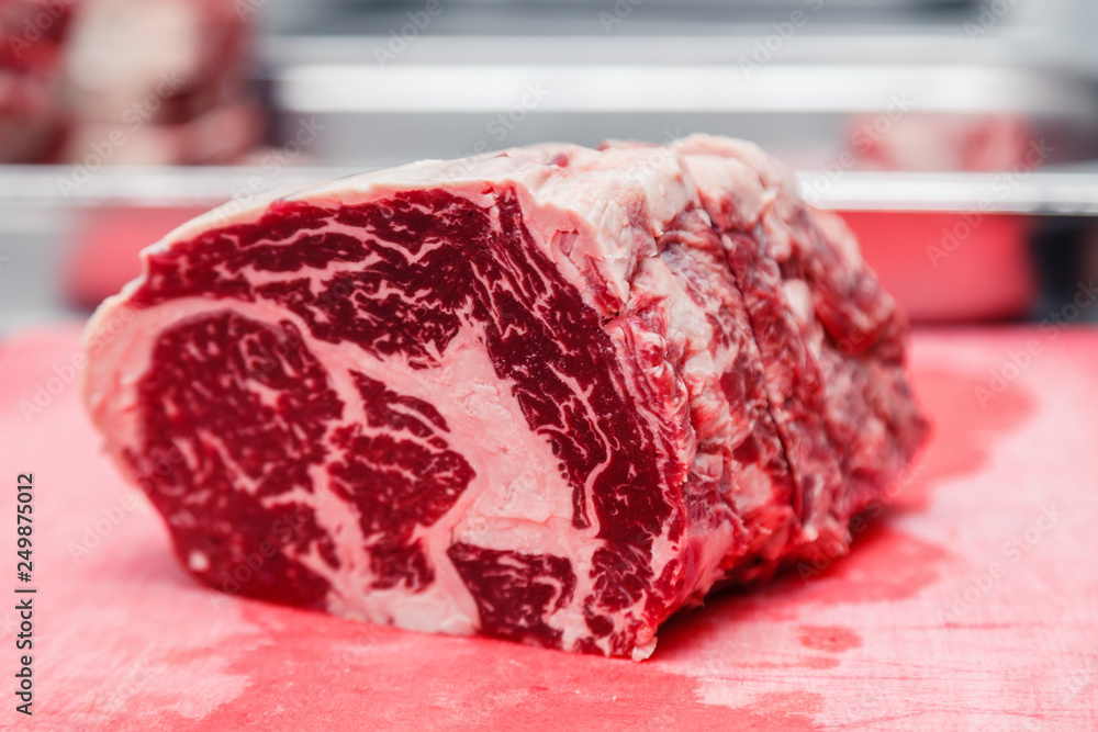 Obraz premium Closeup macro pieces ribeye marbled beef steak on red plastic cutting board on metal table in restaurant kitchen. Concept packing semifinished products, freezing food, steakhouse, grill meat