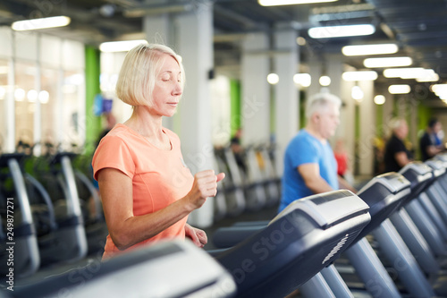 Mature blonde woman in activewear exercising on one of treadmills in contemporary sports center or gym