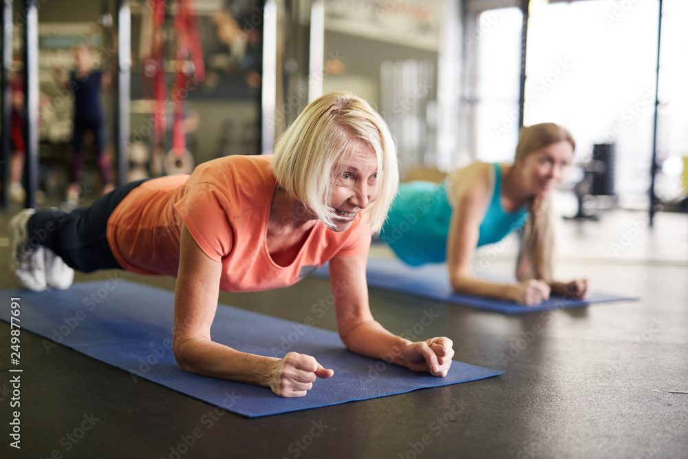 Fototapeta Mature blonde active woman standing in plank on mat during workout in contemporary fitness center