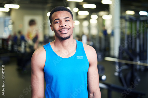 Happy young African-american sportsman or coach in blue vest standing in gym in front of camera