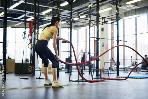 Young active woman with bent knees standing on the floor and exercising with cross training ropes in gym