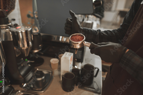 Man barista in an apron is holding a portafilter with coffee and ready for preparing espresso at the coffee shop. Close up photo of bartender with a holder with grinded coffee.