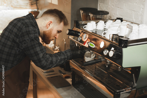 Close up photo of male bearded barista making coffee drink in a restaurant. Young cafe owner is preparing coffee by coffee machine.