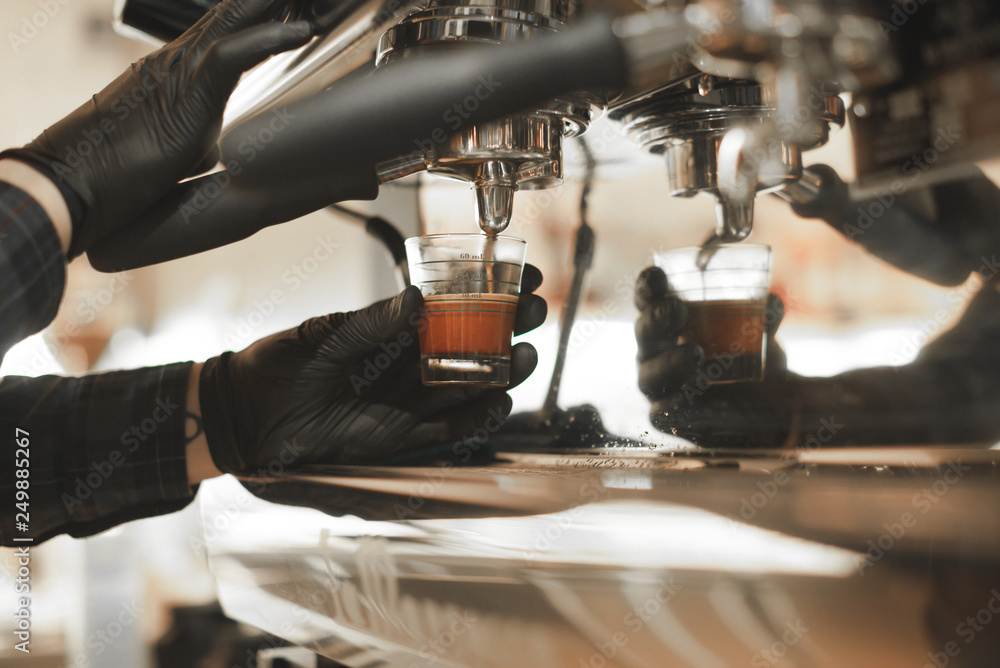Male barista is preparing espresso in a measure glass at the coffee machine. Barista is pouring coffee in a measure glass at the cafe. Close up, selective focus, toned, space for text.