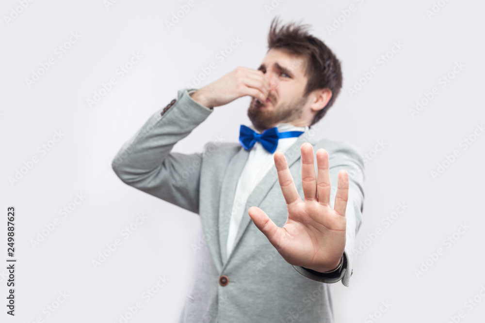 Don't get close. Portrait of handsome bearded man in casual grey suit and blue bow tie standing with blocking ban hand and pinching his nose. indoor studio shot, isolated on light grey background. Don't get close. Portrait of handsome bearded man in casual grey suit and blue bow tie standing with blocking ban hand and pinching his nose. indoor studio shot, isolated on light grey background.