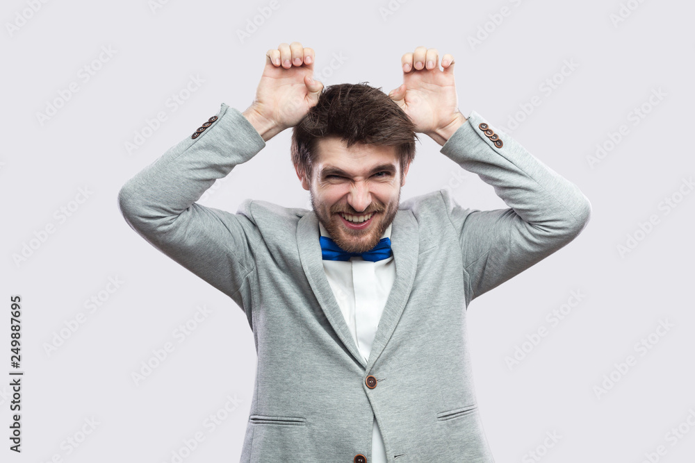 Portrait of funny handsome bearded man in casual grey suit and blue bow tie standing with comic face and bunny ears looking at camera. indoor studio shot, isolated on light grey background. Portrait of funny handsome bearded man in casual grey suit and blue bow tie standing with comic face and bunny ears looking at camera. indoor studio shot, isolated on light grey background.