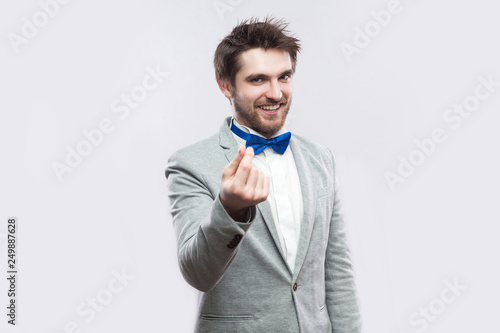 Portrait of funny handsome bearded man in casual grey suit, blue bow tie standing with money gesture hand and looking at camera with cunning face. indoor studio shot, isolated on light grey background