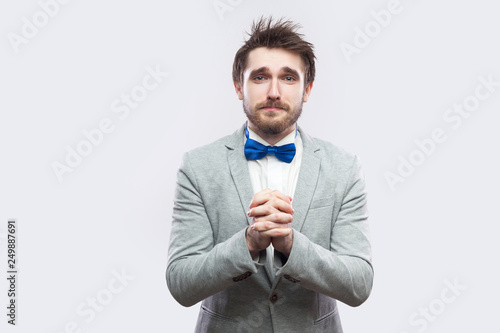 I am sorry, please forgive me. sad handsome bearded man in casual grey suit and blue bow tie looking at camera and asking for help or forgivness. indoor studio shot, isolated on light grey background. photo