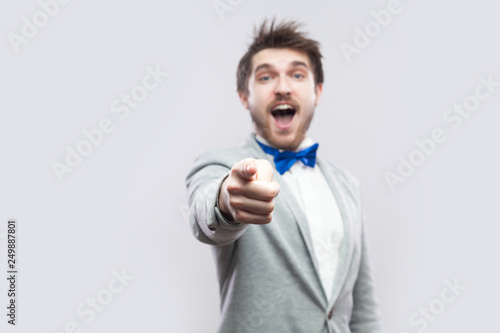 Portrait of surprised handsome bearded man in casual grey suit and blue bow tie standing pointing and looking at camera with amazed face. indoor studio shot, isolated on light grey background.