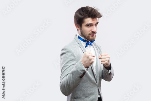 Portrait of serious handsome bearded man in casual grey suit and blue bow tie standing and looking at camera with boxing fists and ready to attack. studio shot, isolated on light grey background.