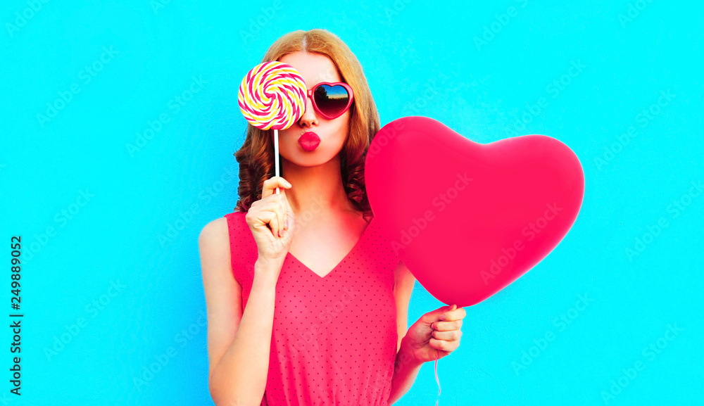 Portrait cool girl with pink heart shaped balloon sending sweet air kiss closing her eye with lollipop on colorful blue background