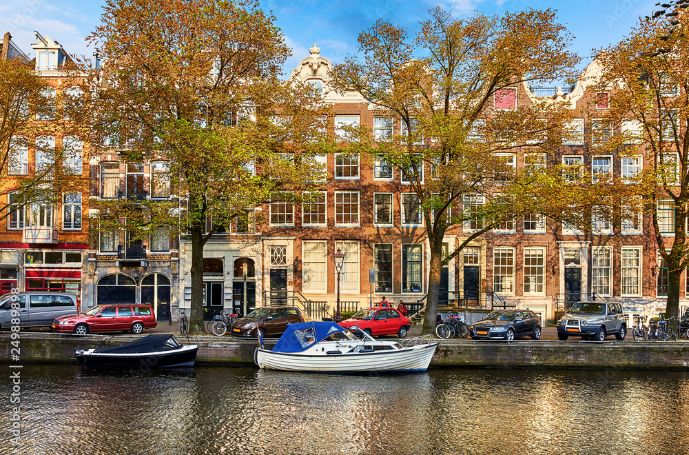 Channel of Amsterdam city. Netherlands. Motorboats by banks on Amstel river among trees and traditional dutch houses above water. Street with parked cars. Sunny spring day with blue sky.