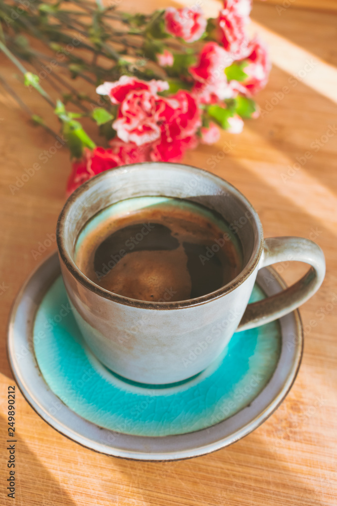 Φωτογραφία Close up view of a blue vintage cup of black coffee and a bouquet of pink carnation flowers on wooden background illuminated by sun rays coming from a side window