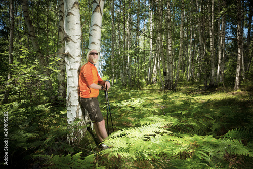 Young man with blonde beard and hiking sticks leaning on a tree in forest