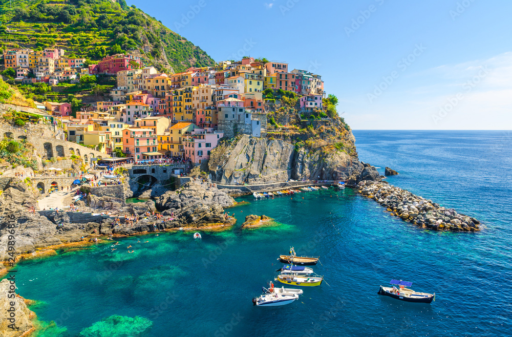 Manarola traditional typical Italian village in National park Cinque Terre, colorful multicolored buildings houses on rock cliff, fishing boats on water, blue sky background, La Spezia, Liguria, Italy