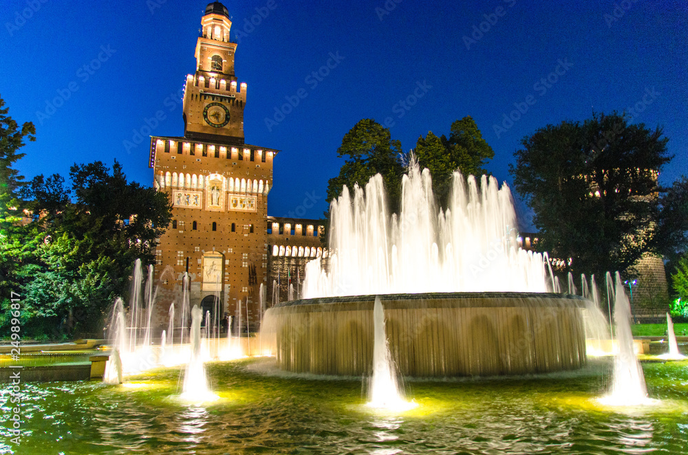 Old medieval Sforza Castle Castello Sforzesco facade, walls and tower La torre del Filarete with lights, trees, lighting fountain at sunset, dusk, twilight, evening, blue sky, Milan, Lombardy, Italy