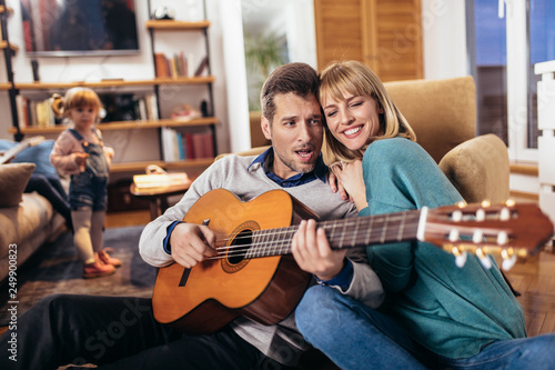 Romantic couple having fun at home, man playing guitar, child in background.