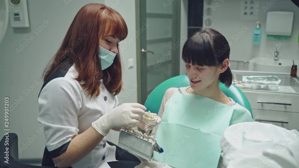 Friendly dentist showing jaw layout to a patient girl sitting in the stomatology clinic. Professional dentist explaining to female patient correct tooth brushing on the dental model Friendly dentist showing jaw layout to a patient girl sitting in the stomatology clinic. Professional dentist explaining to female patient correct tooth brushing on the dental model