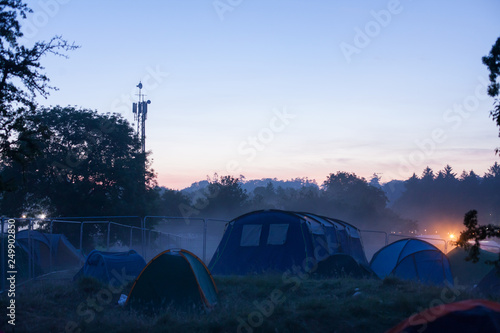 The sun rises behind trees, with the morning mist still visible over tents