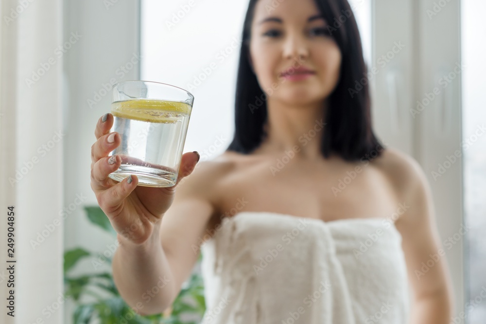 Close-up of glass water with lemon in hand of young woman standing at home near the window. Healthy lifestyle, diet, antioxidant.