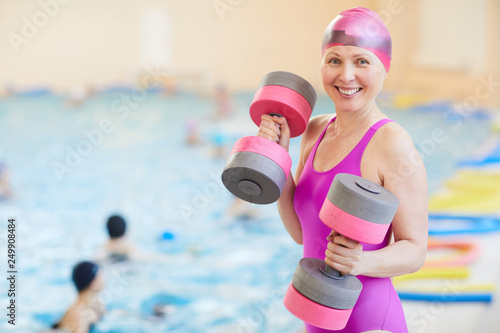 Waist up portrait of active senior woman wearing swimsuit posing by swimming pool holding foam dumbbells and smiling at camera, copy space photo