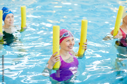 High angle view at group of active senior women exercising in swimming pool, holding pool noodles and smiling, copy space