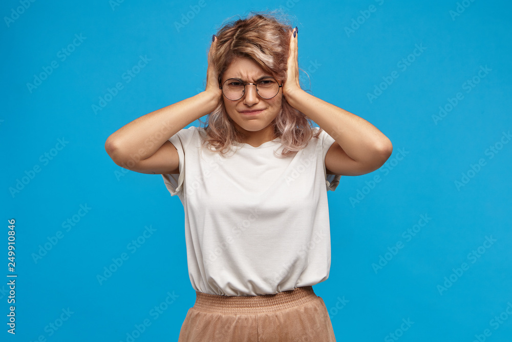 Stop it! Horizontal studio image of frustrated attractive fashionable hipster girl having fight with her jealous boyfriend, can't stand intolerable loud screams, covering her ears, having annoyed look Stop it! Horizontal studio image of frustrated attractive fashionable hipster girl having fight with her jealous boyfriend, can't stand intolerable loud screams, covering her ears, having annoyed look