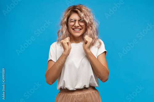 Success, positive news, joy and happiness concept. Ecstatic excited adorable stylish girl in trendy round glasses smiling broadly and clenching fists, glad to receive present on her birthday
