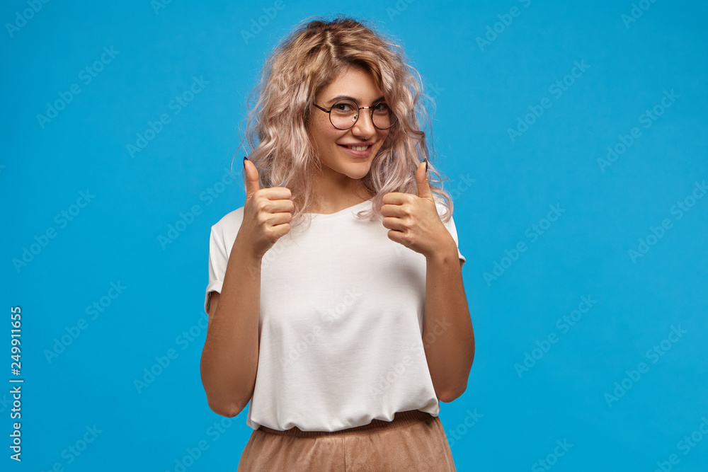 Cheerful hipster girl in trendy round eyewear making thumb up gesture with both hands and smiling joyfully, showing her support and respect to someone, saying Good job, Well done, I like that - obrazy, fototapety, plakaty 