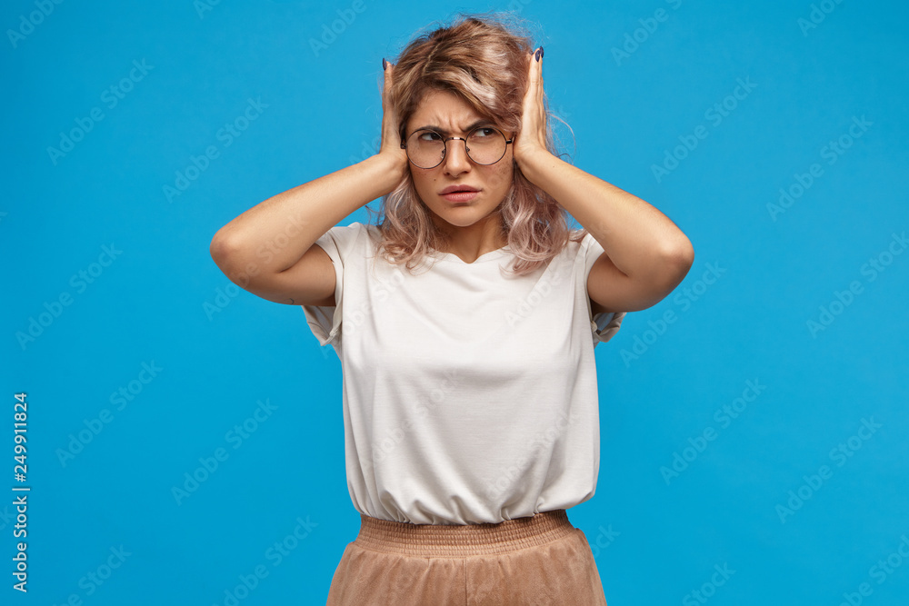 Fototapeta premium Irritated stressed young female in round spectacles covering ears, being disturbed with loud mucis, noisy neighbours and annoying sounds. Body language and negative human facial expressions