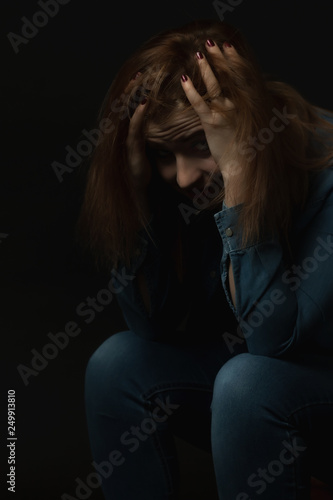 Low key portrait of sitting young woman in trouble holding her head with both hands and looking at the camera. Vertically