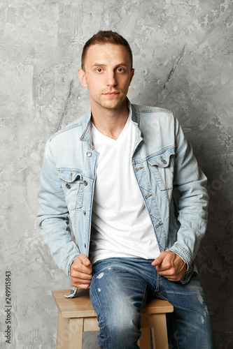 Young handsome guy posing in denim clothes sitting on a chair against the background of a concrete wall