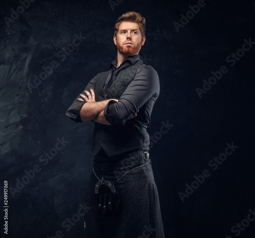Handsome redhead man dressed in an elegant vest with tie and kilt posing with his arms crossed in studio against a dark textured wall
