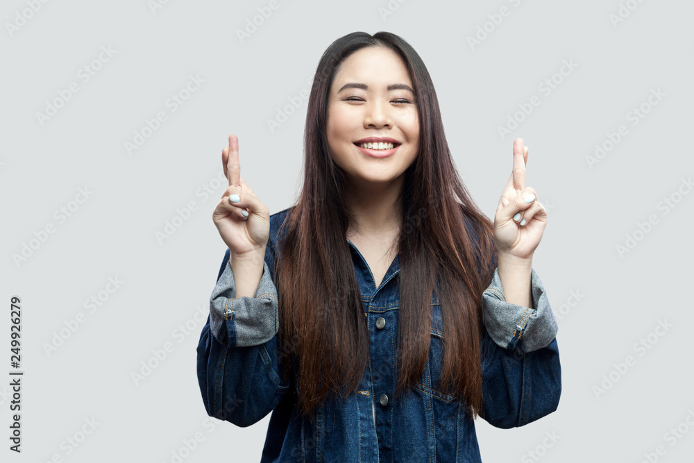 Portrait of hopeful beautiful brunette asian young woman in casual blue denim jacket with makeup standing with crossed finger and praying to win. indoor studio shot, isolated on light grey background. Portrait of hopeful beautiful brunette asian young woman in casual blue denim jacket with makeup standing with crossed finger and praying to win. indoor studio shot, isolated on light grey background.