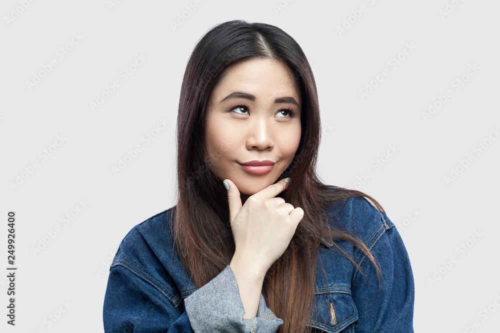 Portrait of dreamy smiley beautiful brunette asian young woman in casual blue denim jacket with makeup standing touching her chin and pondering. indoor studio shot, isolated on light grey background. Portrait of dreamy smiley beautiful brunette asian young woman in casual blue denim jacket with makeup standing touching her chin and pondering. indoor studio shot, isolated on light grey background.