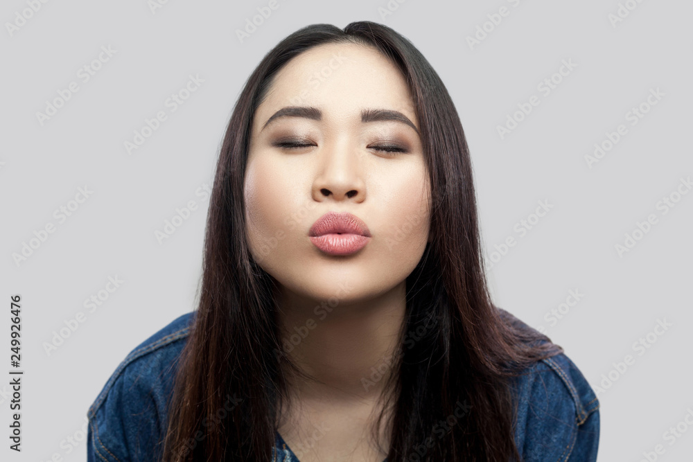Portrait of in love beautiful brunette asian young woman in casual blue denim jacket with makeup standing with closed eyes and sending kiss. indoor studio shot, isolated on light grey background. Portrait of in love beautiful brunette asian young woman in casual blue denim jacket with makeup standing with closed eyes and sending kiss. indoor studio shot, isolated on light grey background.