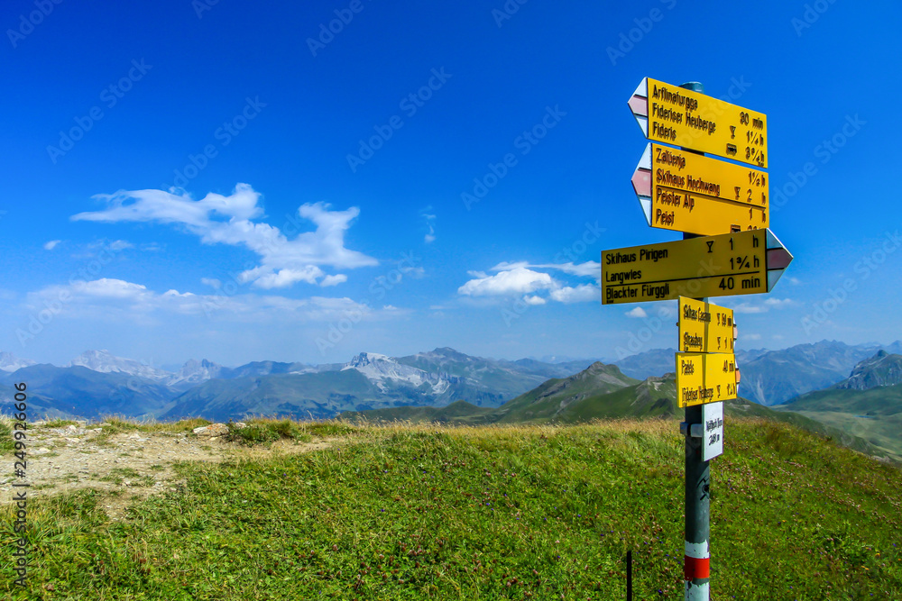 Gelber Wanderwegweiser auf einer gürnen Bergspitze im Engadin in der Schweiz bei blauem Himmel und wenigen weissen Wolken