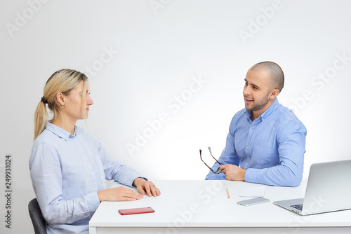 Brutal boss in a blue shirt with glasses interviews new blonde caucasian employee sits by the desk in the office with the white background.