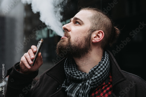 Men with beard hold and smoke his electronic cigarette outdoor in black, grey backgraund
