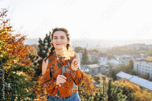 Happy attractive girl holds an autumn maple leaf on sunset panorama of European city background © blackbirdua