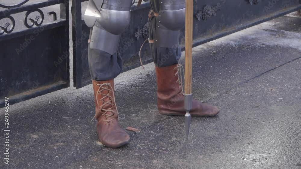 A knight in a metal suit and brown leather boots stands near an iron gate holding a spear in his hand A knight in a metal suit and brown leather boots stands near an iron gate holding a spear in his hand