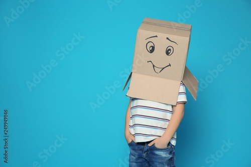Cute little child wearing cardboard box with smiling face on color background. Space for text