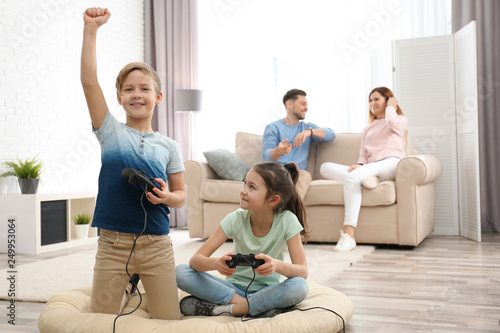 Cute children playing video games while parents resting on sofa at home
