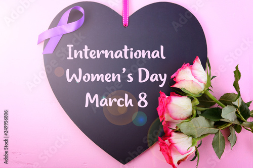 International Women's Day, March 8, heart shaped blackboard greeting with purple ribbon symbol and pink roses on pink wood background with lens flare.