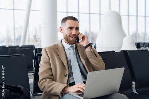 Happy businessman working on the laptop and talking on cellphone at the airport waiting lounge. Handsome caucasian businessman at waiting room in airport terminal © Margo Kos