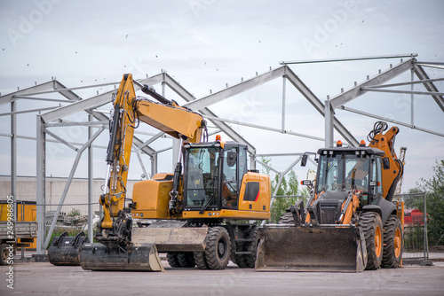 Multiple cars, excavators, trucks, loaders, concrete mixers and construction machinery in large parking lot in industrial territory, next to concrete and asphalt factory  