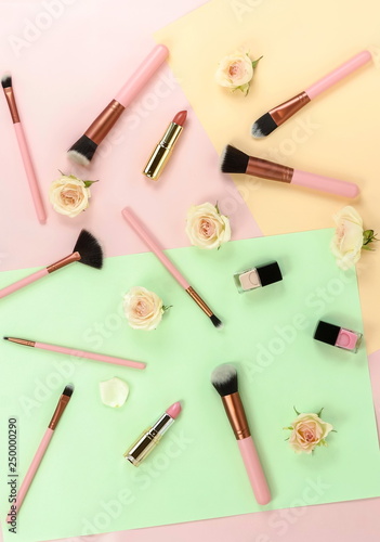 Makeup cosmetic accessories products brushes, lipstick, pink flowers on pastel colors background. Flat lay. Top view. Copy space