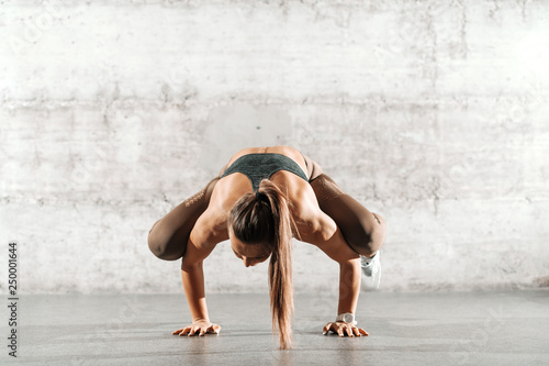 Young sporty brunette with ponytail and in sportswear doing handstand in front of gray rustic wall.